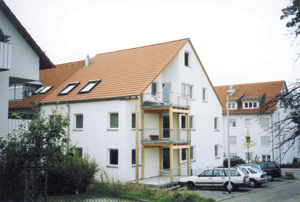 Außenansicht des 1997 erbauten Minimalenergiehauses. Die Holzbalkone sind vom Haus vollständig getrennt. Auf einer Holzschalung befindet sich jeweils eine Dachbahn, die mit einer Gummischnitzelmatte abgedeckt ist, worauf die Gehwegplatten verlegt sind.